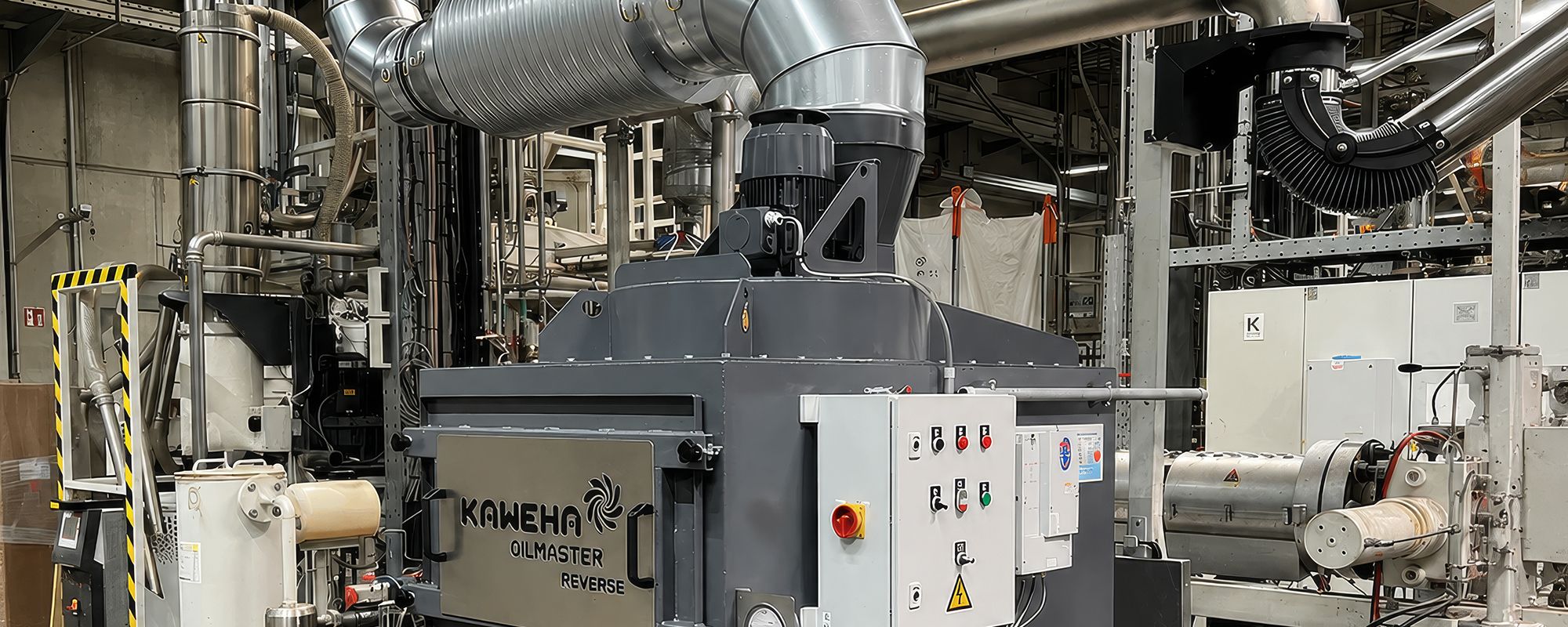 Grijze, kubusvormige KAWEHA OILMASTER REVERSE industriële afzuiginstallatie met bovenaan aangesloten zilveren ventilatiebuizen, in een fabriekshal