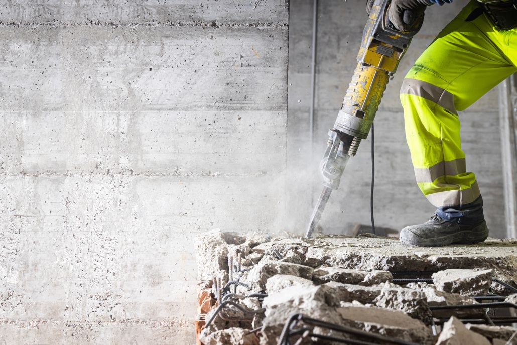 Construction workers wearing high-visibility clothing are using a jackhammer to break up a concrete structure, creating dust in the process