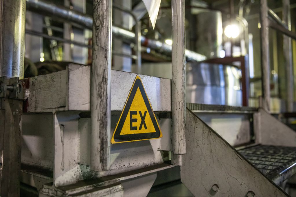 Triángulo de emergencia amarillo sobre un soporte metálico en una planta industrial; al fondo, tuberías y depósitos