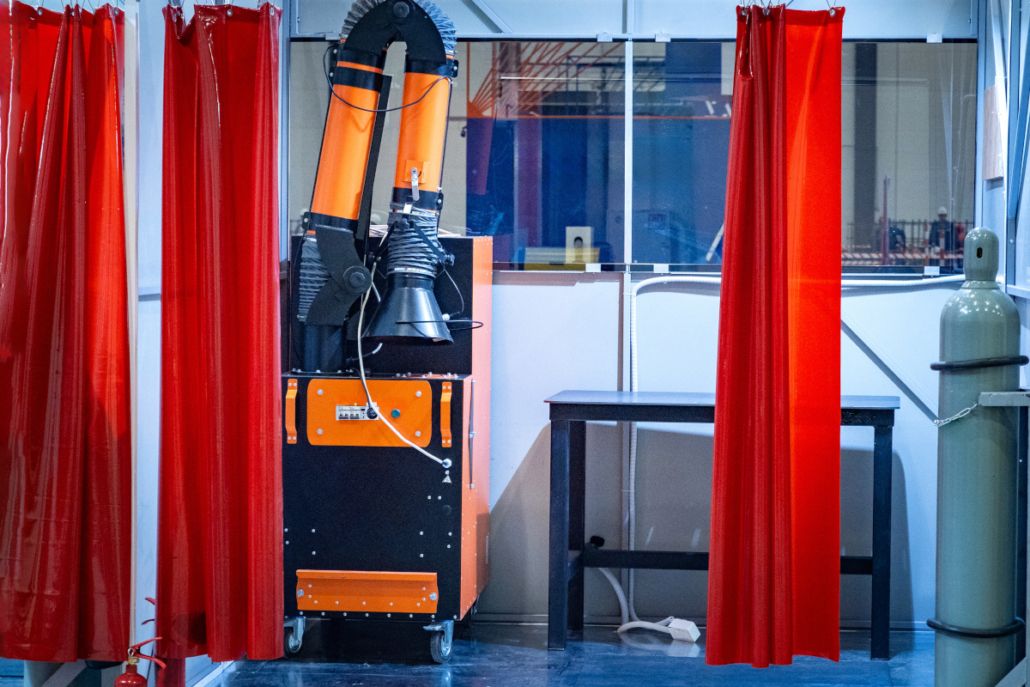 Una escena industrial con un aspirador de color naranja y una bombona de gas gris en una zona de trabajo. Unas cortinas de color rojo brillante enmarcan la zona