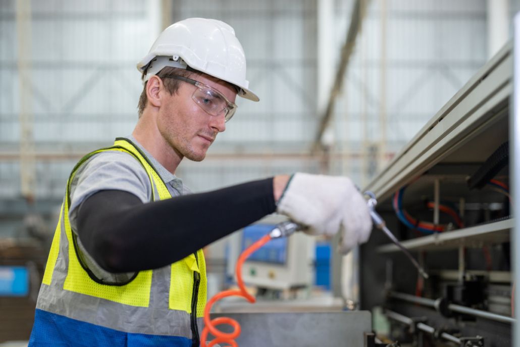 Techniker mit Schutzhelm, Schutzbrille und Warnweste bei Arbeiten an einer Industriemaschine, mit Druckluftwerkzeug in der Hand