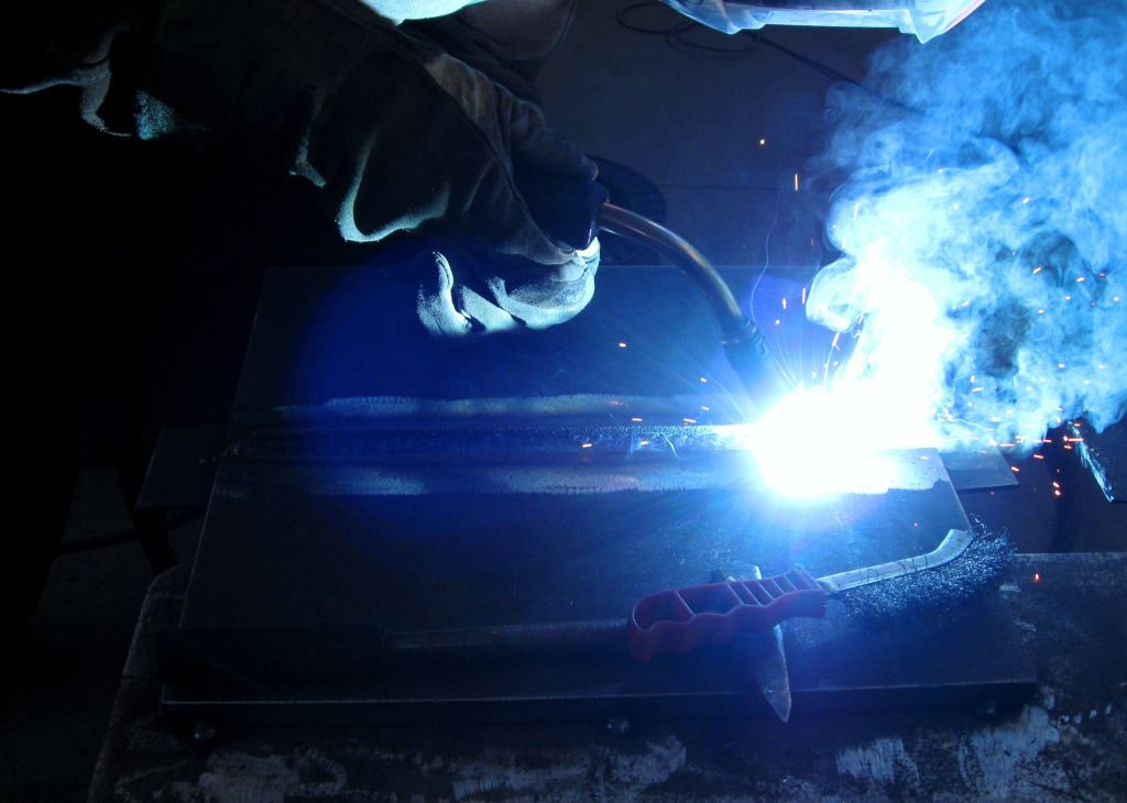 Schweißer mit Schutzhandschuhen beim Schweißen eines Werkstücks, dabei entstehender Schweißrauch, Funken und heller Lichtbogen